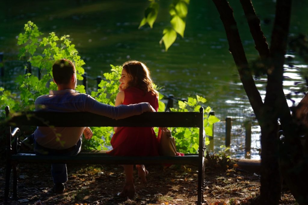A woman considers a man as they sit on a bench in front of a lake